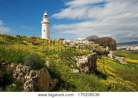 White old lighthouse over meadow covered by grass yellow flowers and rocks against the background of scenic sky
