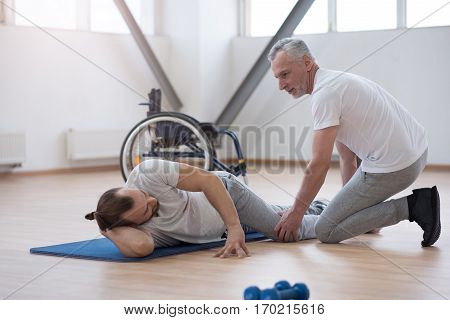 Enjoying helping people. Cheerful helpful skilled physical therapist stretching the disabled man and assisting while holding leg of the patient and expressing joyfulness