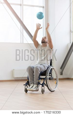 Amusing myself. Cheerful amused young handicapped sitting in the wheelchair in the gym while exercising and playing with the ball