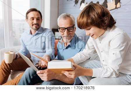 Look at this. Cheerful positive nice child sitting next to his grandfather and showing him a page in the book while reading it