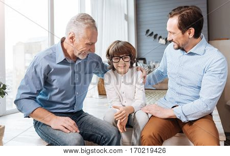 Bad eyesight. Cute pleasant young child sitting between his father and grandfather and wearing glasses while having problems with the eyesight