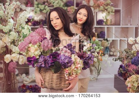 Beautiful asian florist girls holding bouquet and basket of flowers for sale against floral bokeh background in flower shop indoors. Two attractive asian females florists working in retail store. 2 playful fashion models in tender dresses posing smiling a
