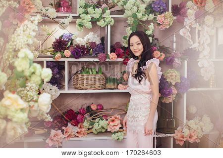 Beautiful girl in tender pink dress standing against floral background in flower shop. Joyful asian female florist with alot of flowers . Playful fashion model looking at camera.