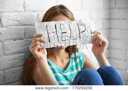 Sad teenage girl sitting near brick wall and holding paper with word HELP