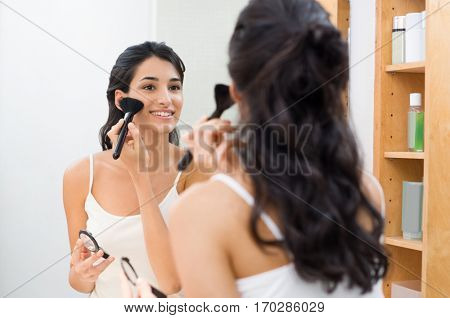 Beautiful young woman doing make up in her bathroom. Happy young woman applying blush on cheek in front of a mirror in bathroom. Brunette girl applying foundation on face with brush.