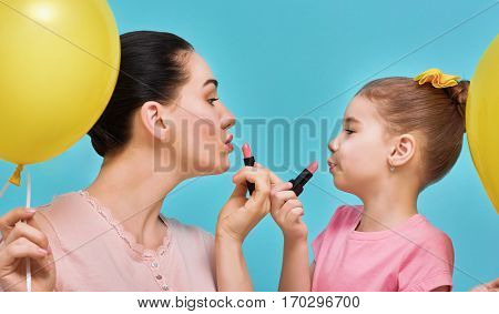 Funny family on a background of bright blue wall. Mother and her child girl are doing your makeup and having fun. Mom and daughter wear lipstick. Yellow, pink and turquoise colors.