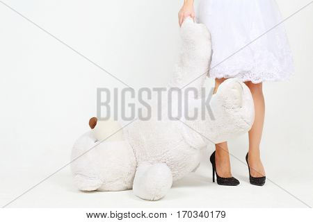 Woman in white dress holds legs of big toy bear in white studio, noface