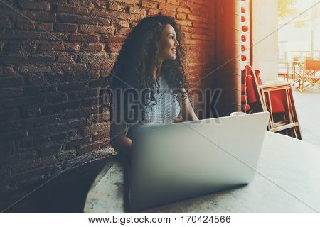 Portrait of curly smiling hipster girl using laptop computer in cafe charming caucasian brunette women looking aside while working on her net-book