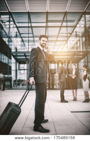 Businessman holding suitcase talking on mobile phone in office premises