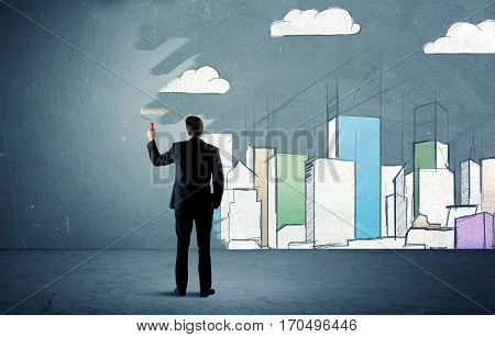 An elegant businessman standing with a paint roller transforming a blue wall into urban city landscape drawing sketch including skyscrapers and clouds concept