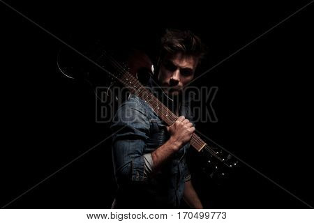 dramatic young guitarist looking back while holding guitar on shoulder, on black studio background
