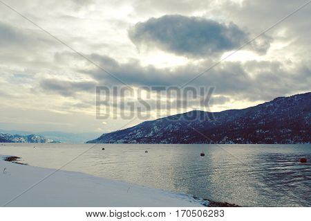 Winter landscape view of lake and mountains with sunshine through clouds over mountain. Sunlight on calm lake with snow covered rocks and shoreline.