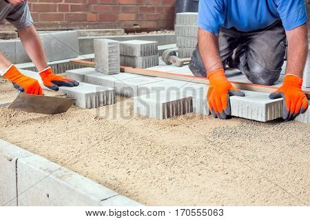 Hands of two builders laying paving stones on a prepared levelled sand base placing them into position around a new house