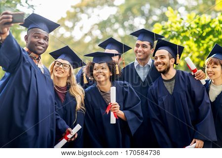 education, graduation, technology and people concept - group of happy international students in mortar boards and bachelor gowns with diplomas taking selfie by smartphone outdoors
