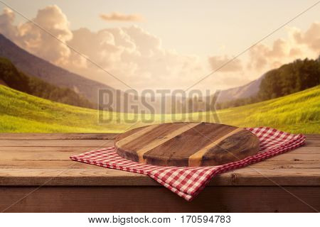Wooden cutting board with checked tablecloth on table over beautiful landscape background