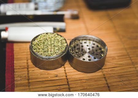 Steel or metal grinder full of freshly ground cannibus flower. Related smoking accessories nearby.