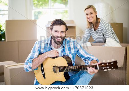 Man playing a guitar while woman unpackaging cardboard boxes in background in their new house