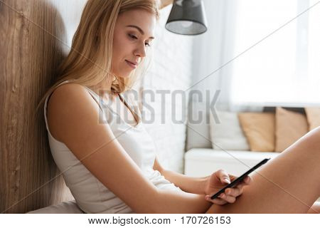 Side view of woman lying on bed with smartphone with window on background