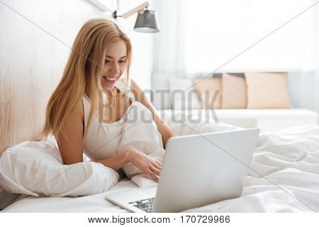 Smiling woman using laptop and lying on bed in bedroom