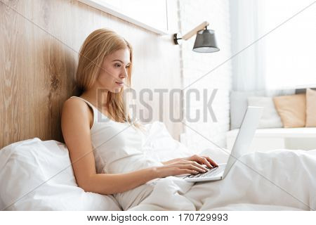 Side view of concentrated woman lying on bed in bedroom with laptop