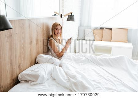 Side view of pretty woman lying on bed with smartphone in hands and looking at camera