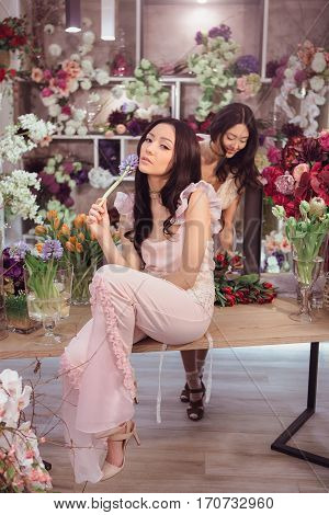 Fashion models in tender dresses posing and looking at camera. Beautiful asian florist girls with flowers in hands against floral bokeh background in flower shop indoors. Two attractive asian females florists working in store.