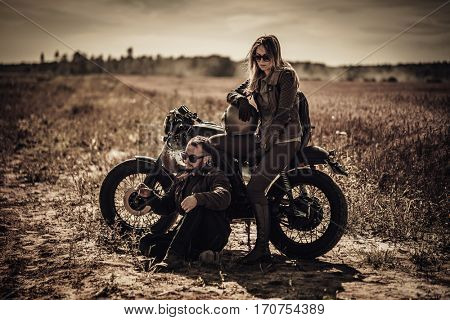 Young, stylish cafe racer couple on vintage custom motorcycles in field.