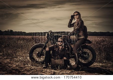 Young, stylish cafe racer couple on vintage custom motorcycles in field.