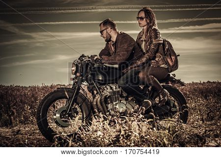 Young, stylish cafe racer couple on vintage custom motorcycles in field.