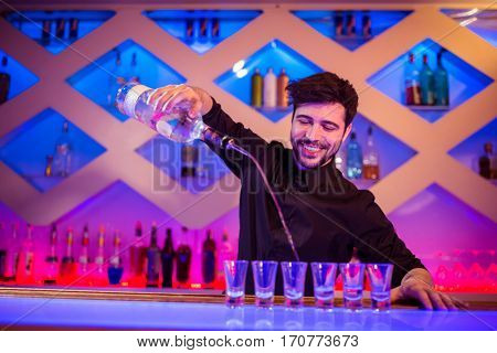 Handsome bartender smiling while pouring alcohol in shot glass at bar counter