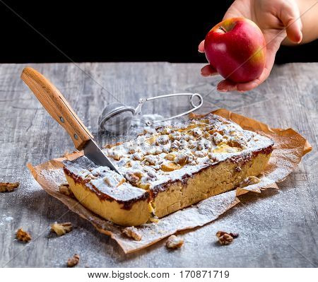 Apple pie dusted with powdered sugar on a grey background. The knife stuck into the tart. Red Apple in hand. Selective focus