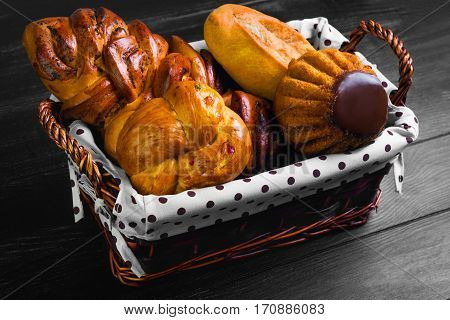 Assortment of fresh pastries buns bread - large loaf Pie with poppy seeds buns with candied fruit cupcake bun with chocolate in special wicker basket for pastries bread. Black wooden background.