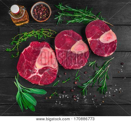 Raw meat beef veal shank slices meat for Osso Buco cooking on dark black wooden background. Ingredients for beef meat Osso Buco sage leaves thyme rosemary pepper salt pepper vegetable oil. Top view.