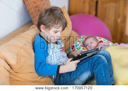 Preschool kid boy playing games tablet computer during babysitting. Cute newborn baby looking on brother. Adorable kids and children at home.
