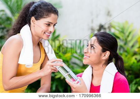 Cheerful young woman giving a bottle of natural plain water to her friend during workout session