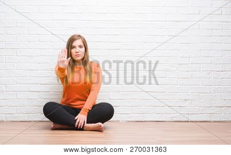 Young adult woman sitting on the floor in autumn over white brick wall with open hand doing stop sign with serious and confident expression, defense gesture