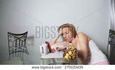Hungry Sad Woman Sits In A Living Room At A Table At Night, She Eats A Liver And Uses Her Smartphone