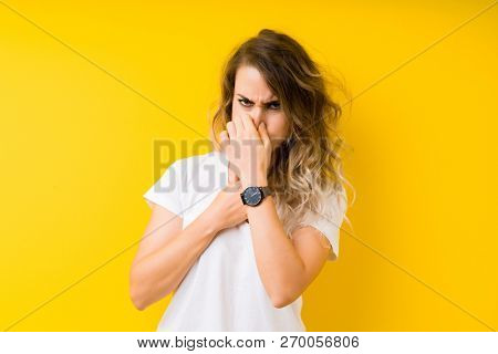 Young beautiful blonde woman over yellow background smelling something stinky and disgusting, intolerable smell, holding breath with fingers on nose. Bad smells concept.