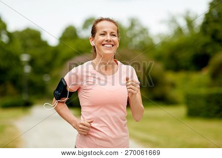 fitness, sport and healthy lifestyle concept - smiling woman with earphones wearing armband for smartphone, jogging at summer park and listening to music