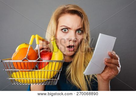 Shocked Woman Holding Shopping Basket With Fruits Looking At Bill Receipt Being Scared Of Huge Price