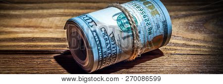 A Roll Of Dollars On A Wooden Textured Table In The Dark Illuminated By A Ray Of Light.