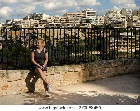 Young Blond Girl In Gray Dress With Pink Backpack Siting On The Stone Gate