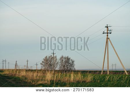 Atmospheric Landscape With Power Lines In Green Field With Dirt Road On Background Of Trees Under Bl
