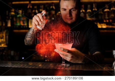 Barman With Tattoos And Beard Sprinkling Red Colored Bitter Into A Cocktail Glass Against Dark Backg