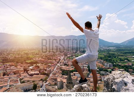 Man Standing On The Top Of The Mountain With His Hands Up Looking At Beautiful Sunset And Cityscape.