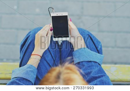 A Young Pretty Student Looks At The Phone And Listens To Music In Headphones Sitting On A Bench In T