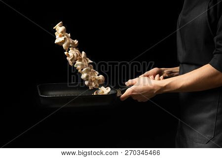 Professional Cook. The Chef Prepares A Dish With Champignon Mushrooms In A Saucepan. On A Black Back