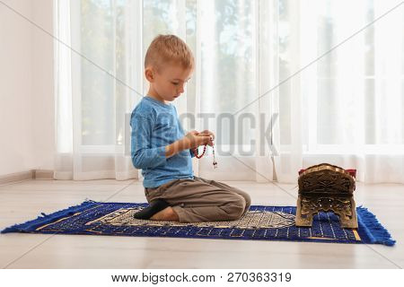Little Muslim Boy With Misbaha And Koran Praying On Rug Indoors