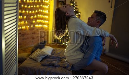 Young Lovers Are In A Hug During Lovemaking On Background Of The Bed, Christmas Tree And Decorations