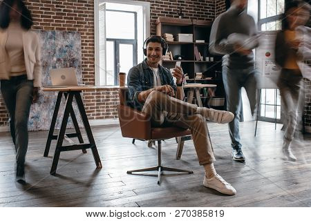 Casual Businessman In Heaphones Sitting On Chair With Colleagues In Motion Blur In Modern Loft Offic